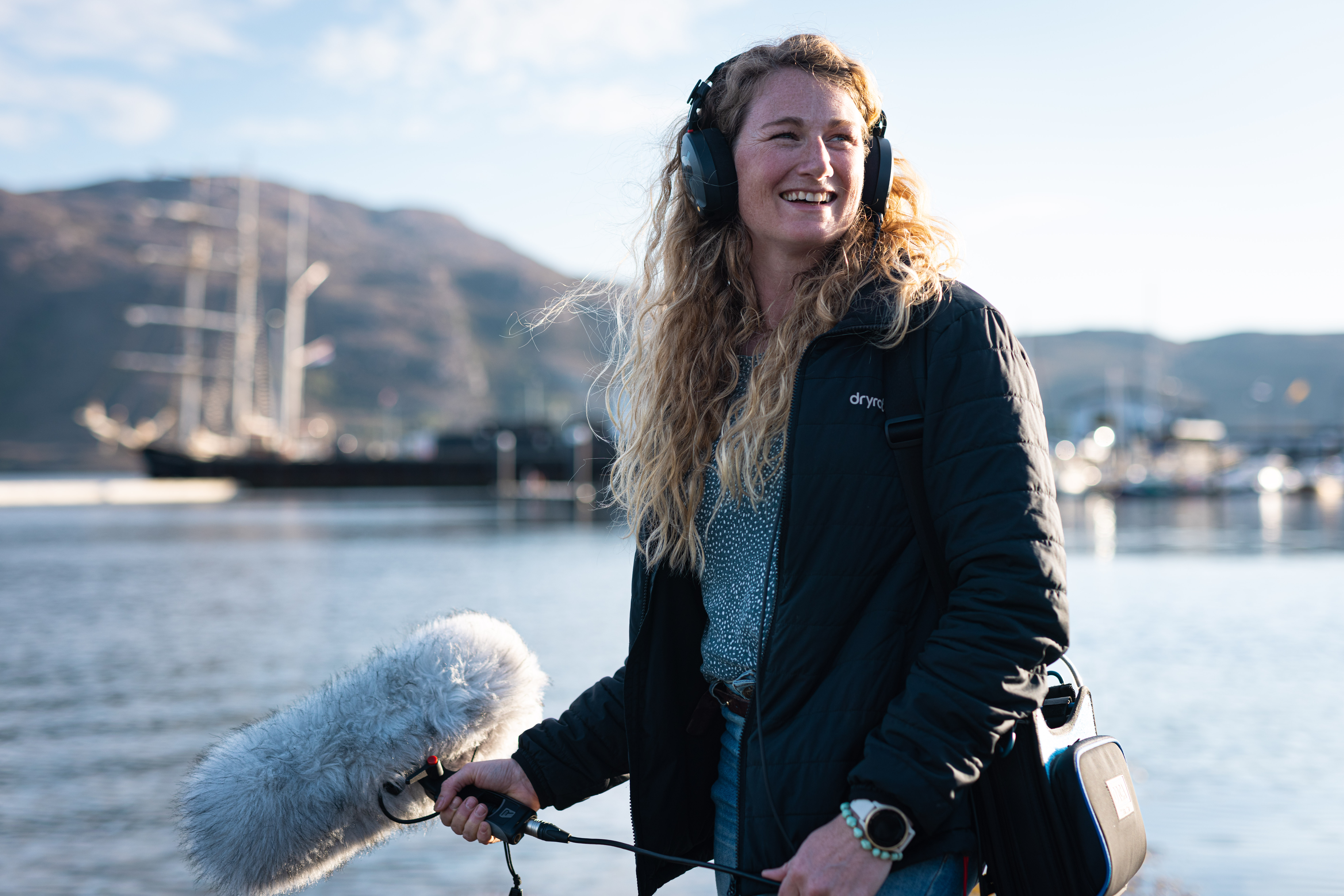 Cal Major holding a podcast mic, smiling looking past the camera from the sea shore