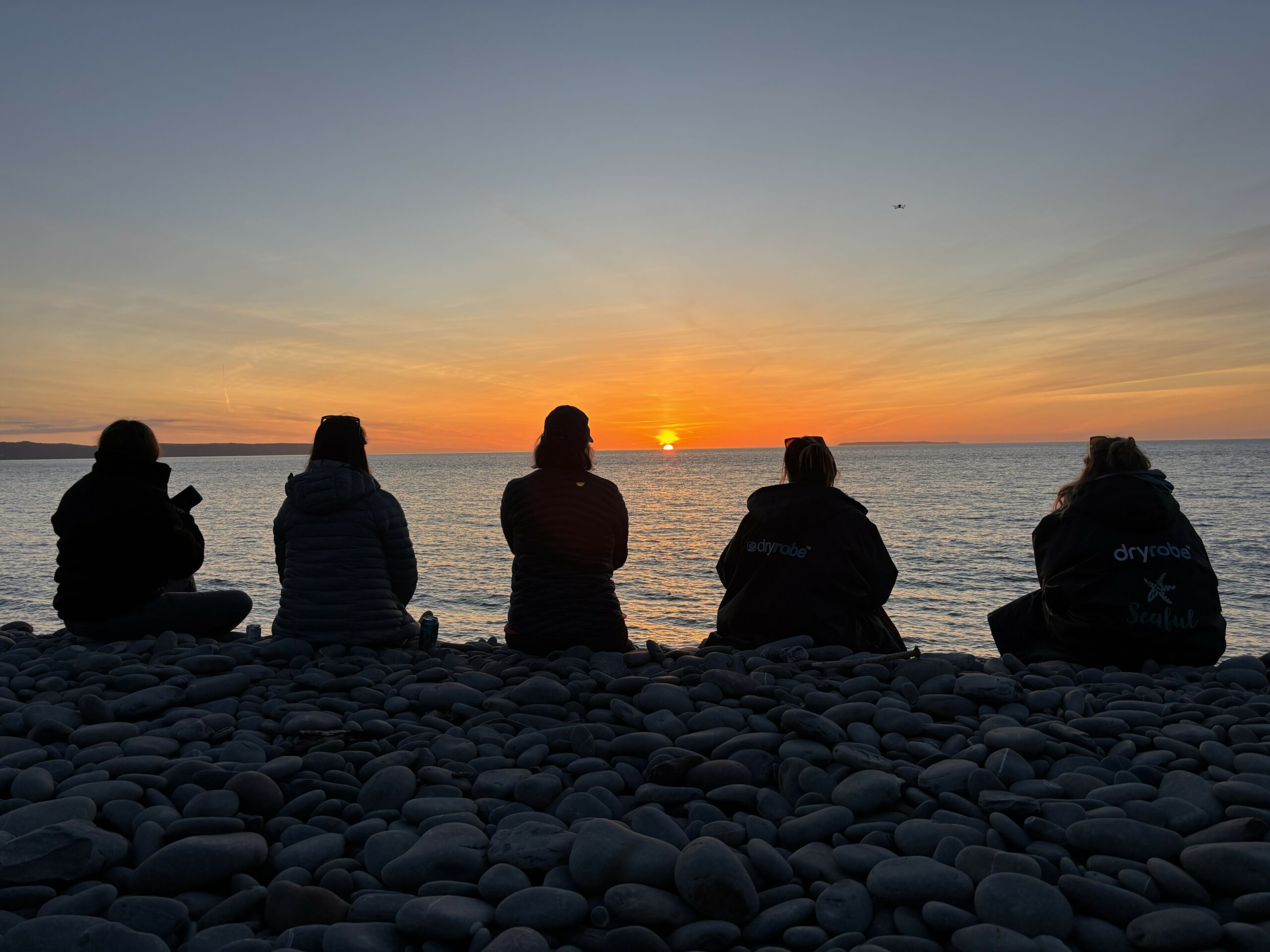 Five people sit on a pebble beach watching the sunset over the sea