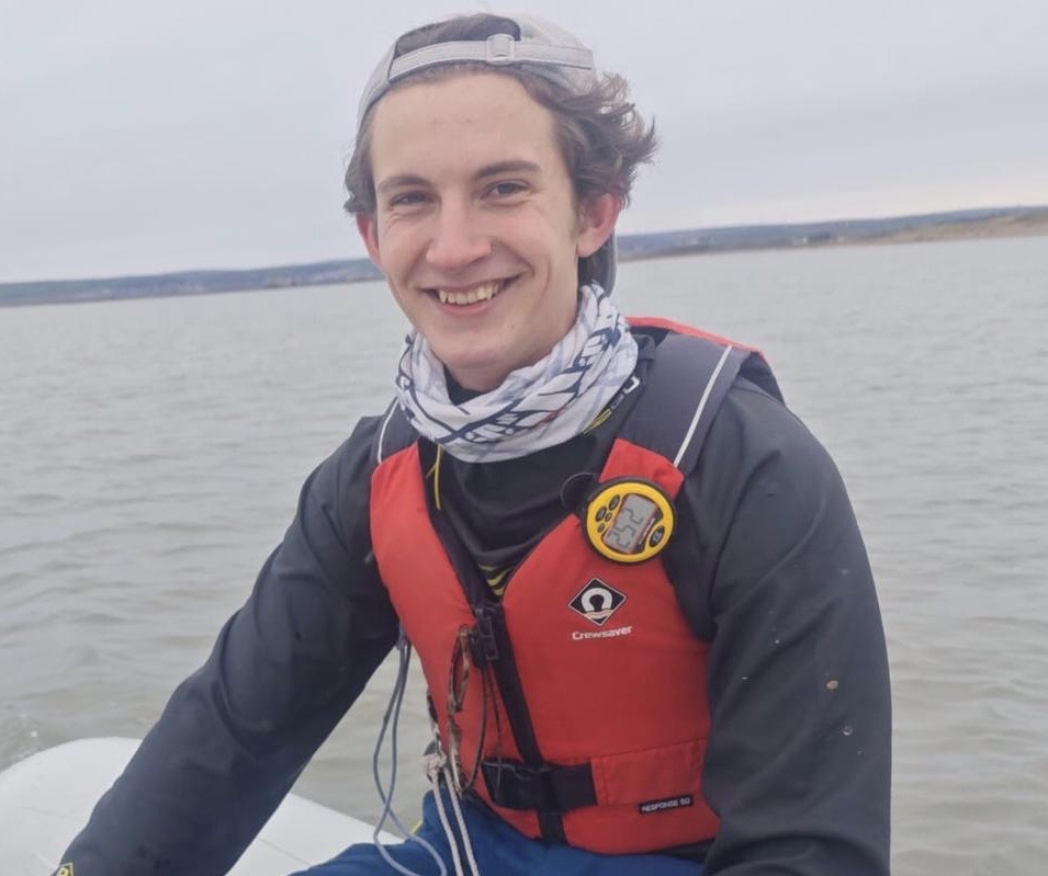 A young man wearing a buoyancy aid, jacket, scarf and cap sits on the edge of a boat