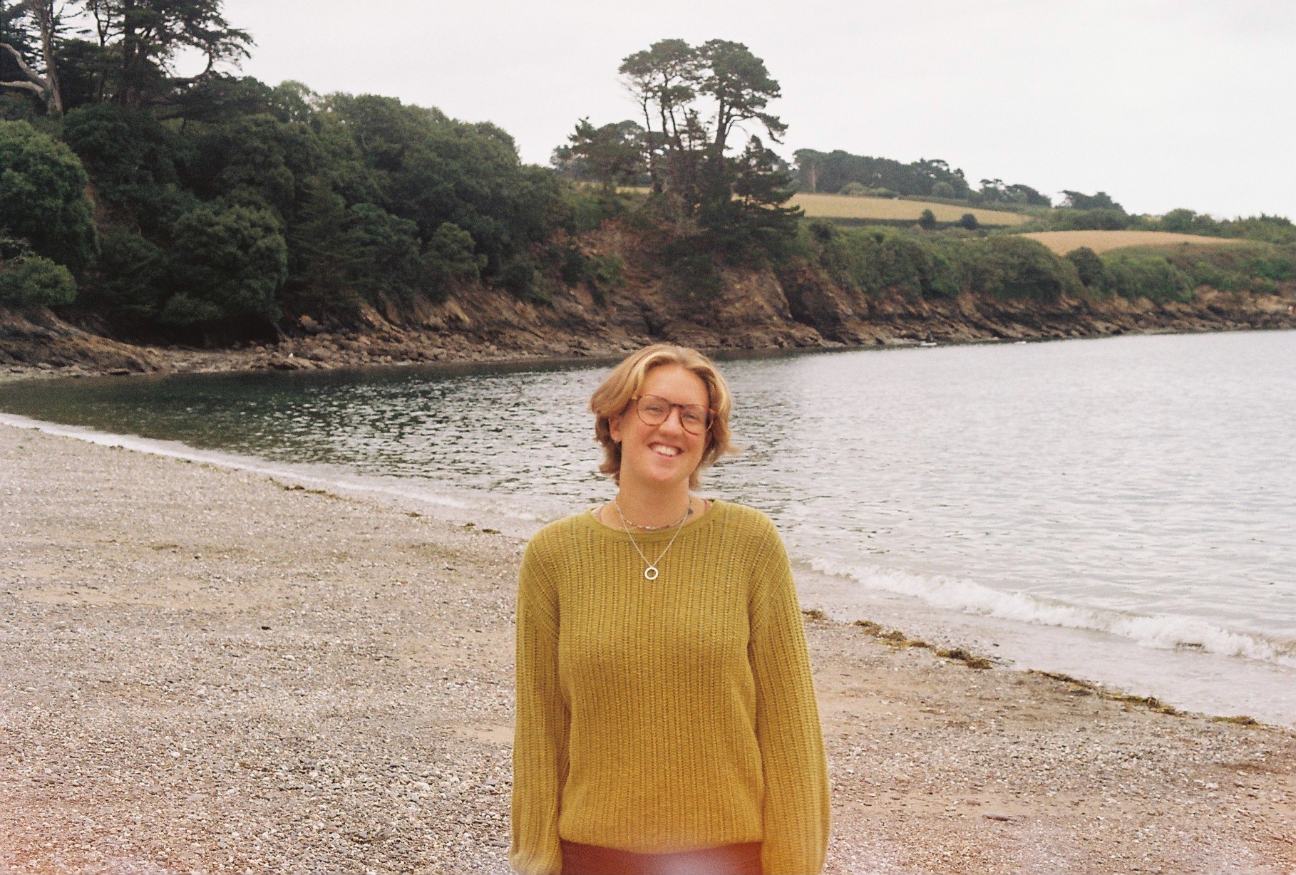 A woman wearing a yellow jumper stands on a beach with the sea and plants in the background
