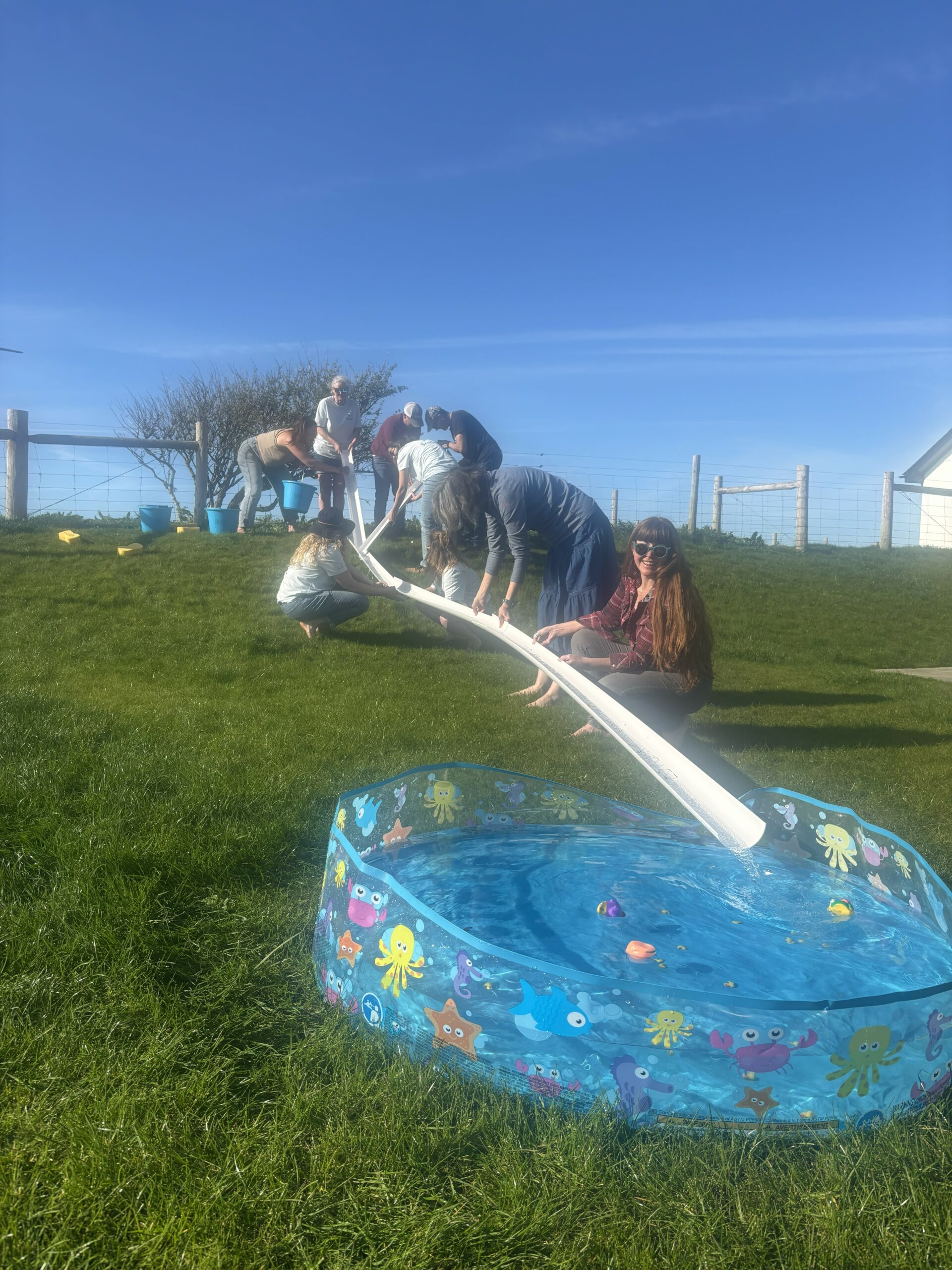 A group of adults hold up a piece of white piping that is draining water into a paddling pool